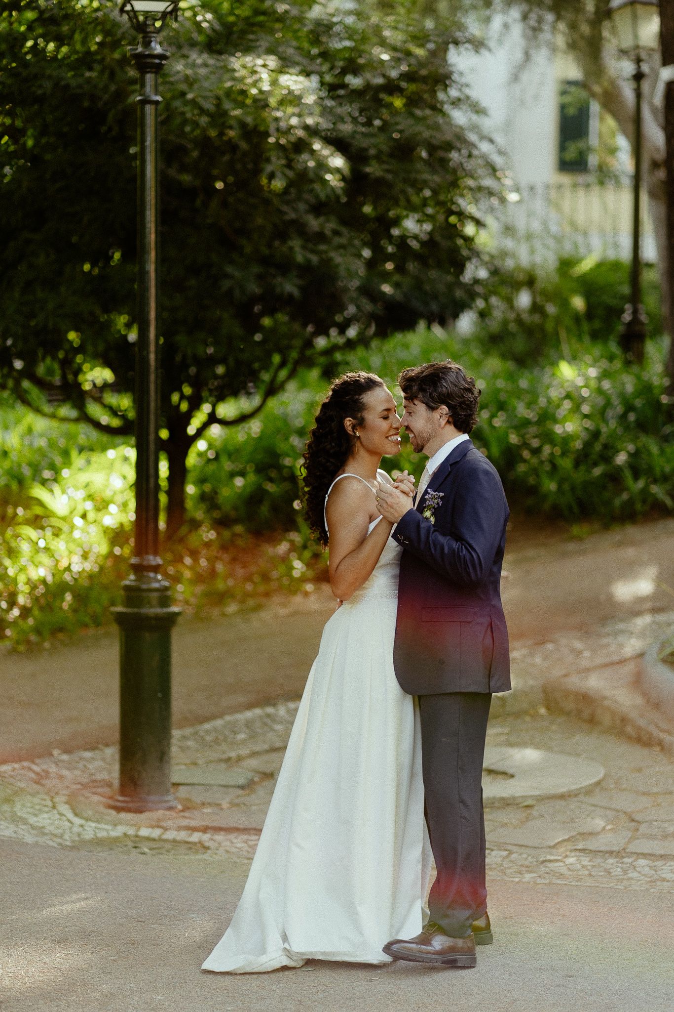 A bride and groom share a romantic dance outdoors near a lamppost surrounded by lush green trees on their wedding day.