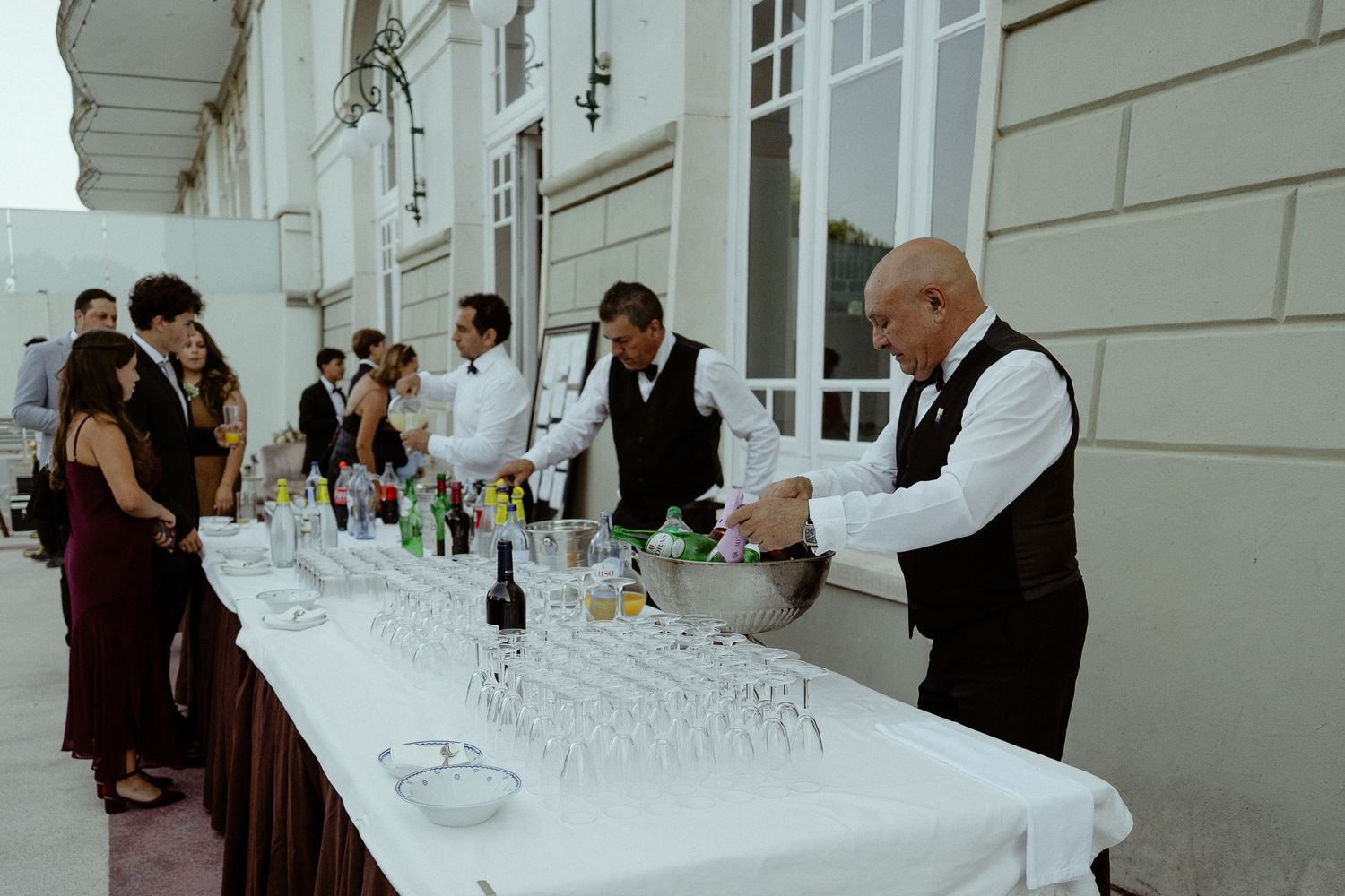 Bartenders in vests serve drinks at an elegant outdoor wedding reception bar with champagne glasses.