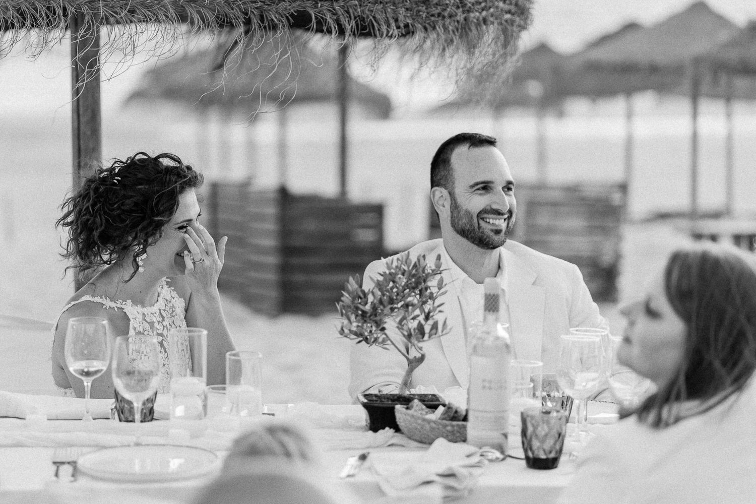 Couple laughing at outdoor beach wedding reception dinner under thatched umbrellas in black and white photo.