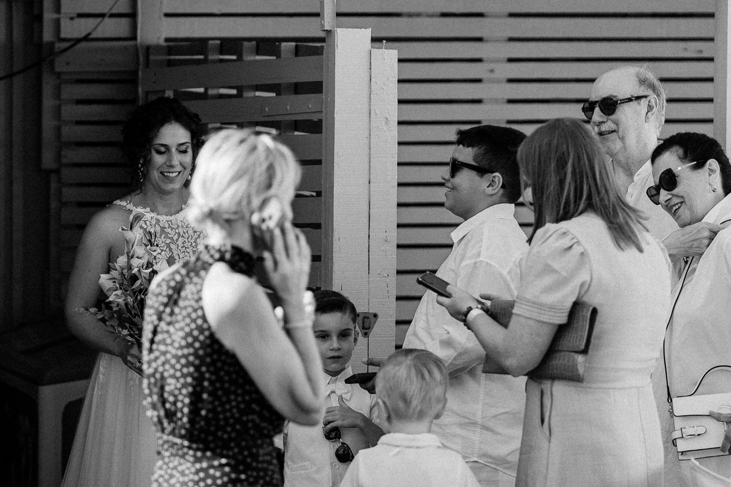 Black and white wedding photo of guests socializing outdoors, bride holding bouquet, children and adults in elegant attire.