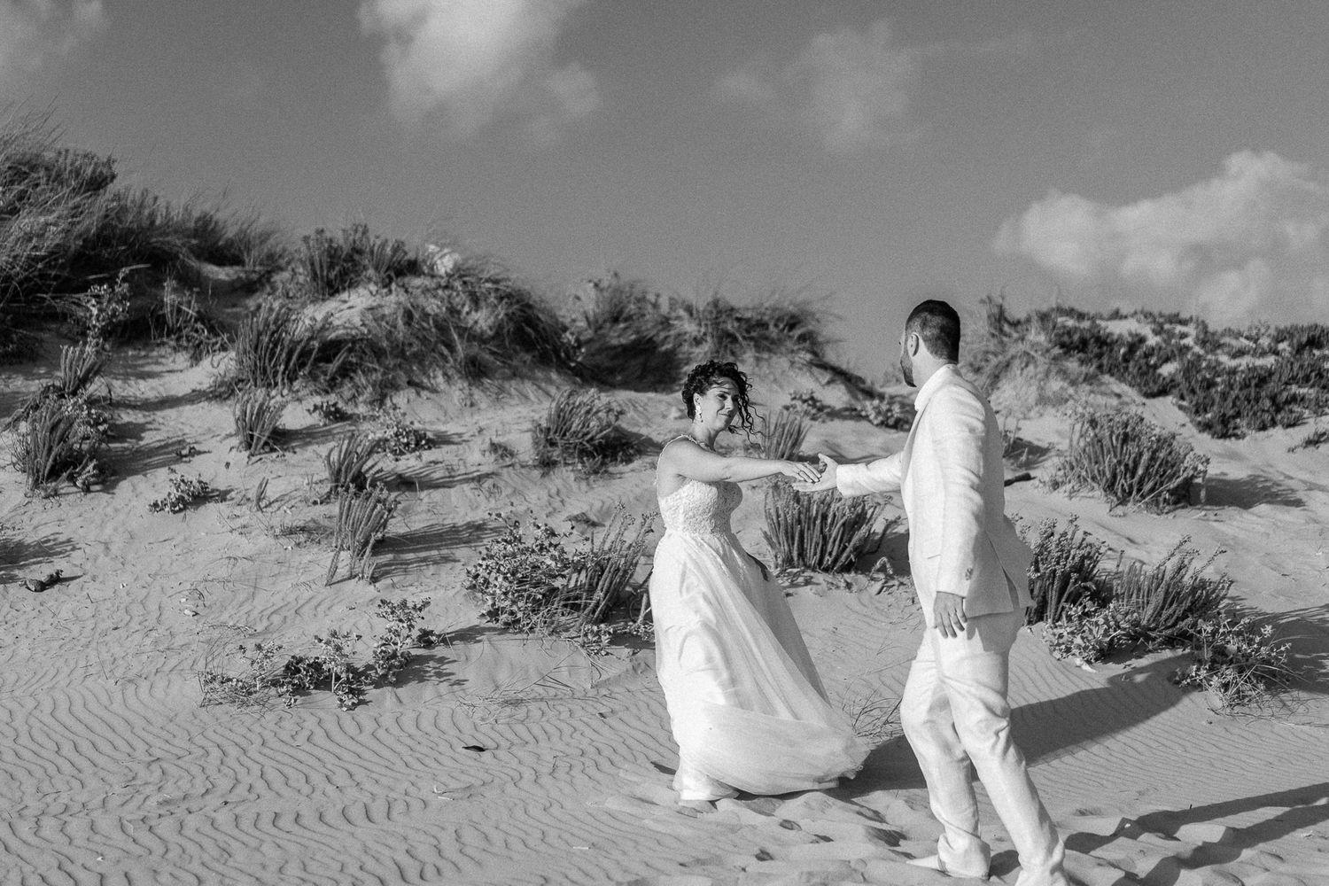Black and white photo of a bride and groom holding hands on sandy beach dunes with grass and cloudy sky.