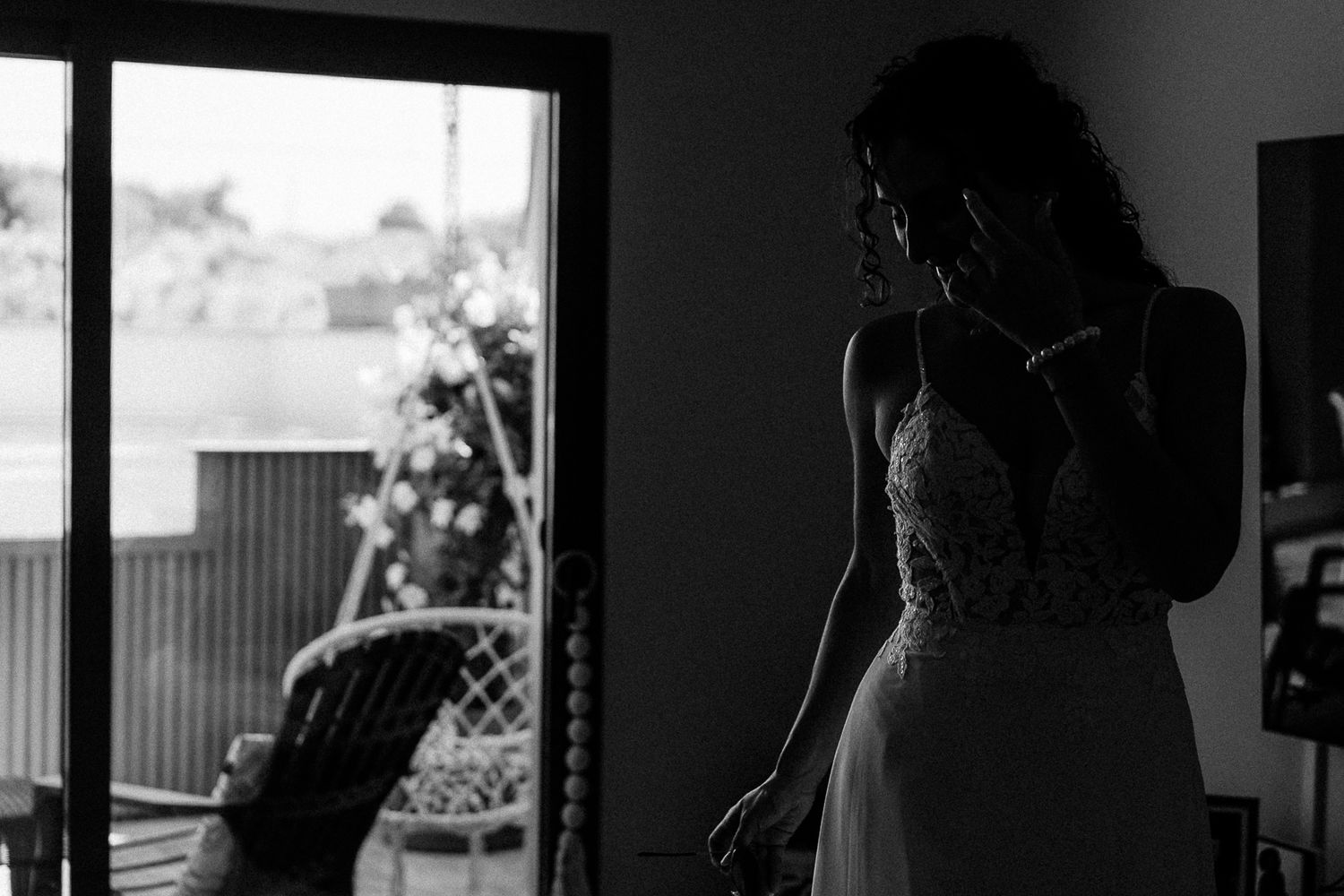 Black and white silhouette of a bride in a lace wedding dress standing near a window on her wedding day.