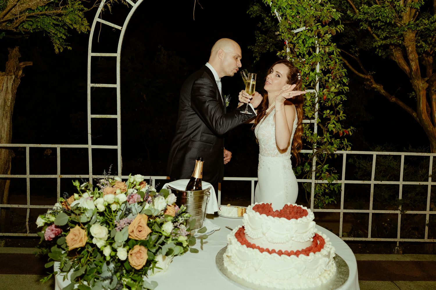 Bride and groom toasting with champagne beside a white wedding cake adorned with strawberries at an outdoor reception.