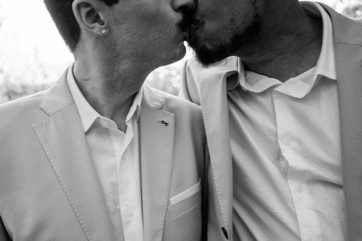 Black and white photo of two men in light suits sharing a kiss at a wedding ceremony.