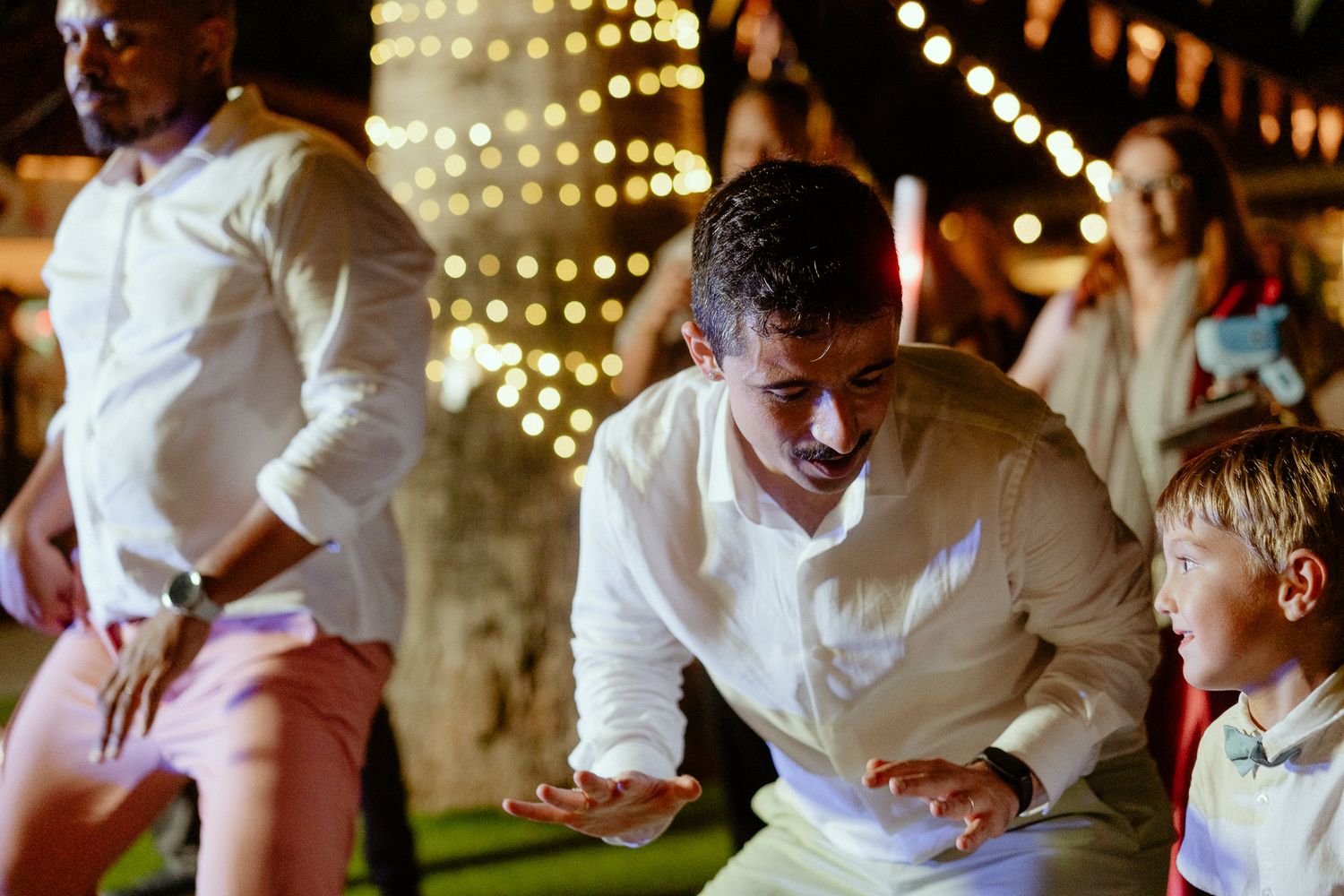 Man in white shirt dancing energetically at an outdoor evening wedding reception with string lights in background.