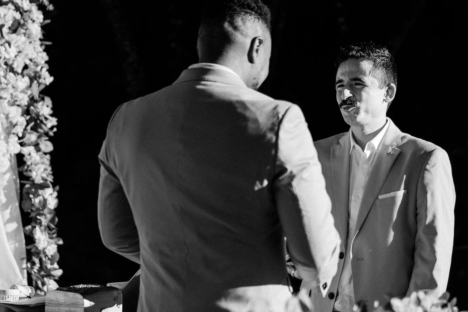 Two men in suits share a joyful moment at a wedding ceremony, smiling and holding hands in black and white.