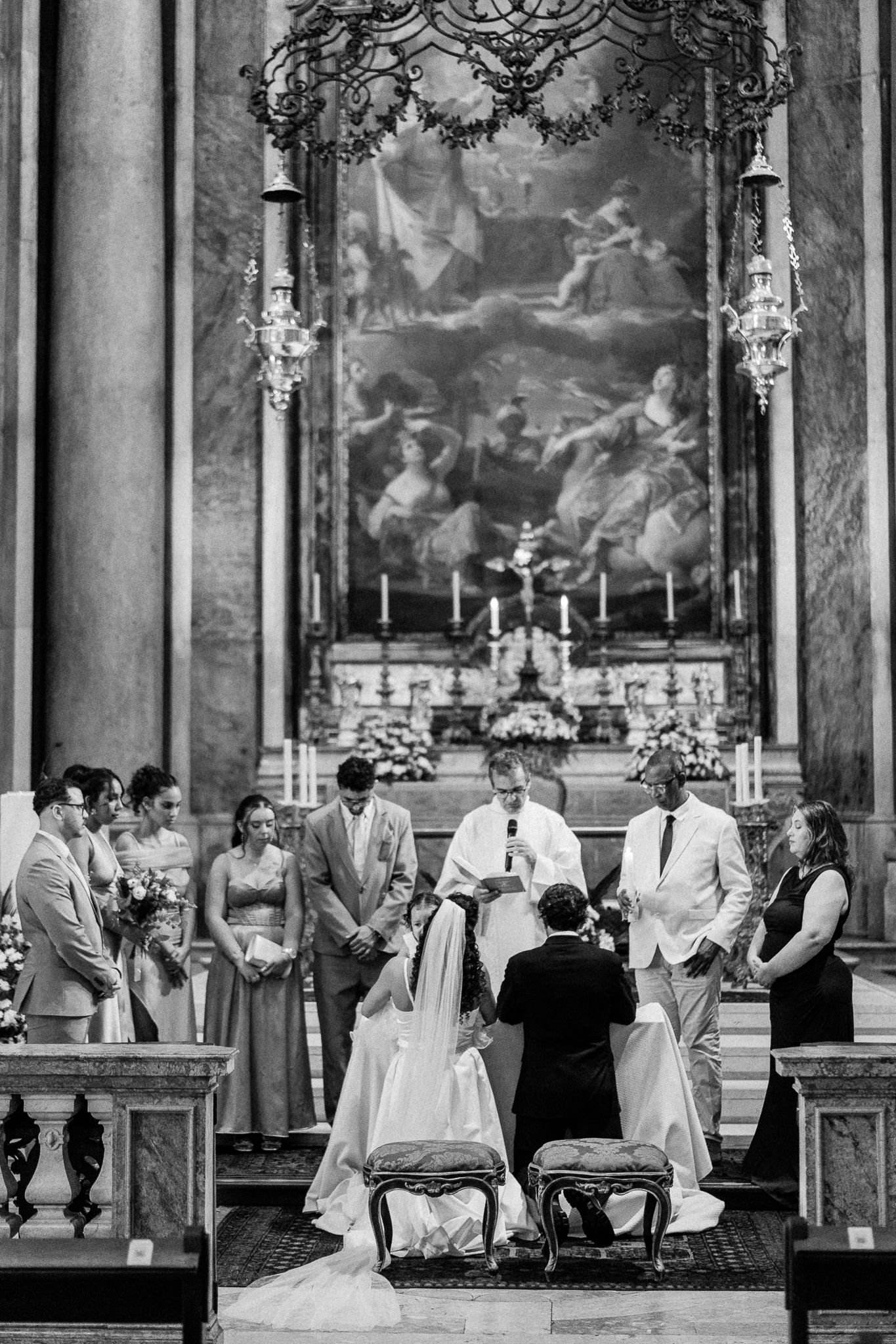 Black and white photo of a wedding ceremony at an ornate church altar with bridal party, officiant, and large painting backdrop.