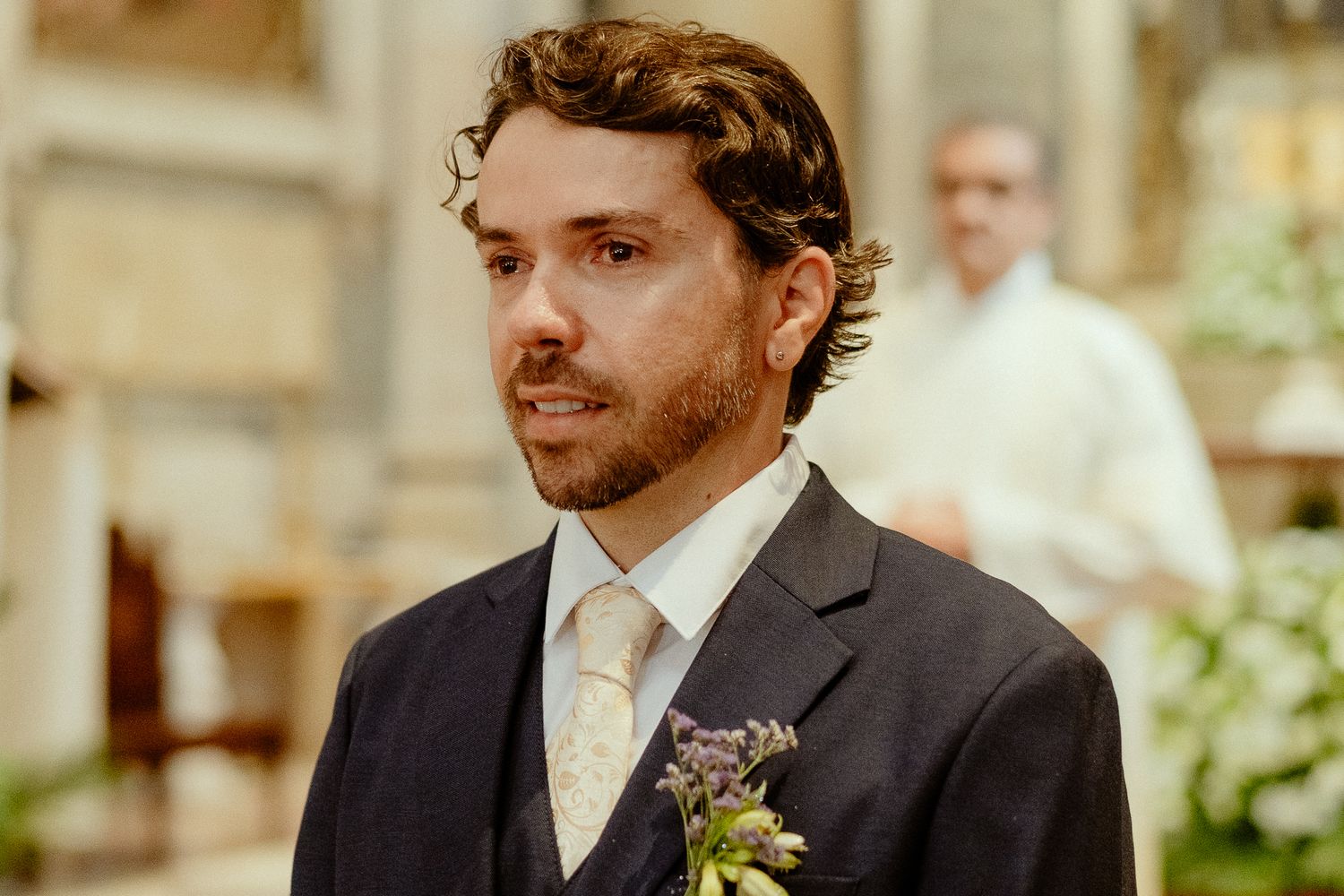 Groom in dark suit with floral boutonniere standing at church altar during wedding ceremony.