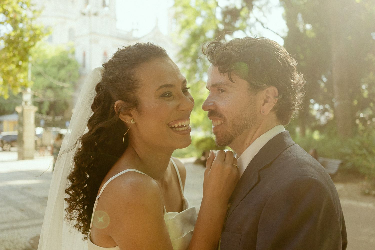 Joyful interracial newlywed couple smiling outdoors on their wedding day, bride in veil and groom in suit.