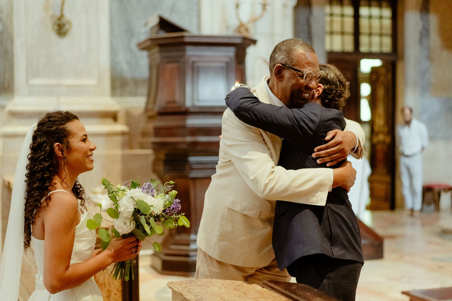 Bride holding bouquet watches as groom embraces guest in elegant church during wedding ceremony.