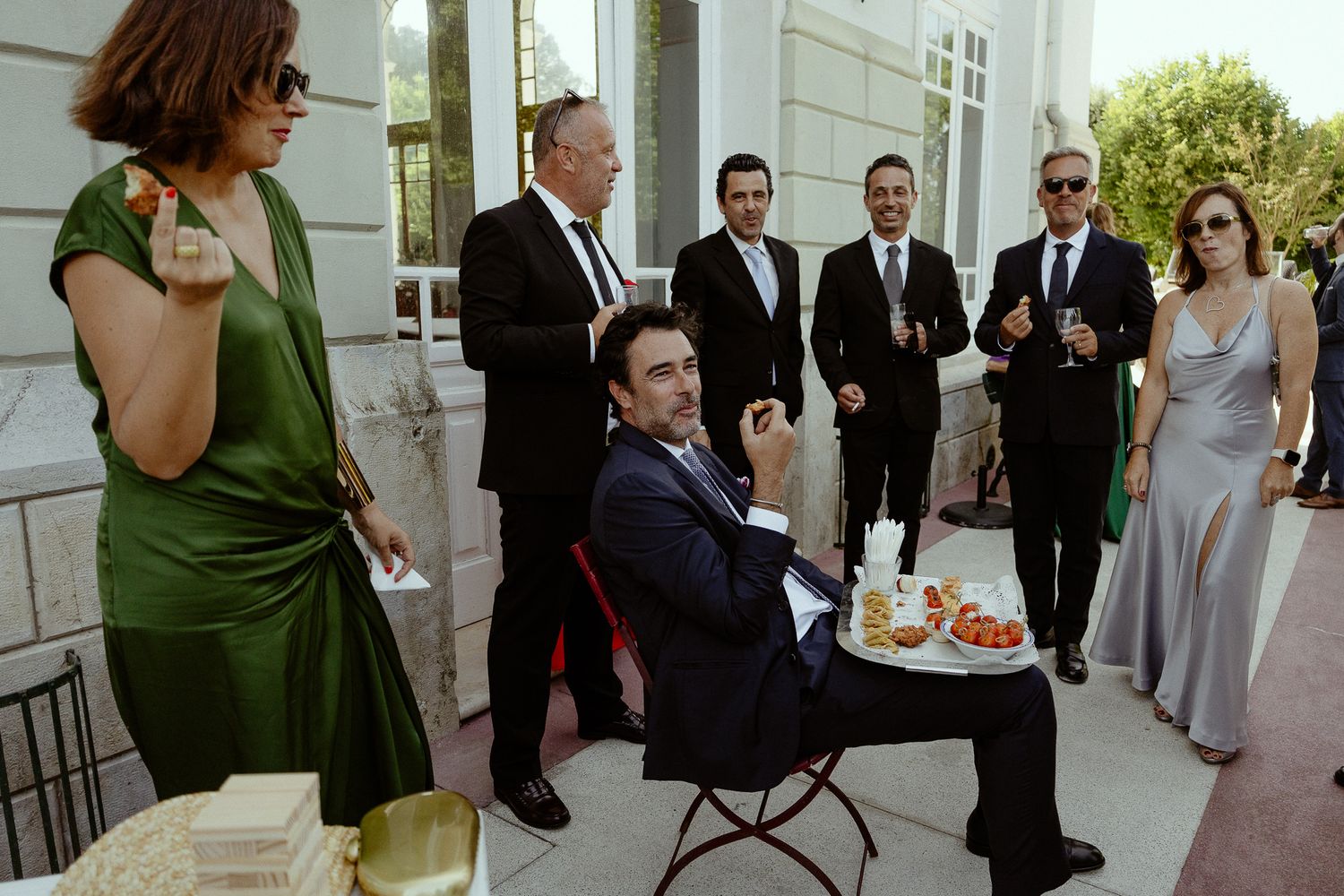 Formally dressed guests socialize at an outdoor event, with one man seated at a table with refreshments.