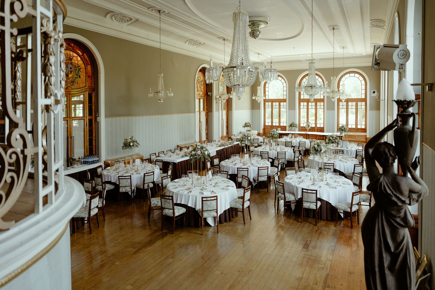Elegant ballroom with round tables, white linens, chandeliers, arched windows, and hardwood floors for weddings.
