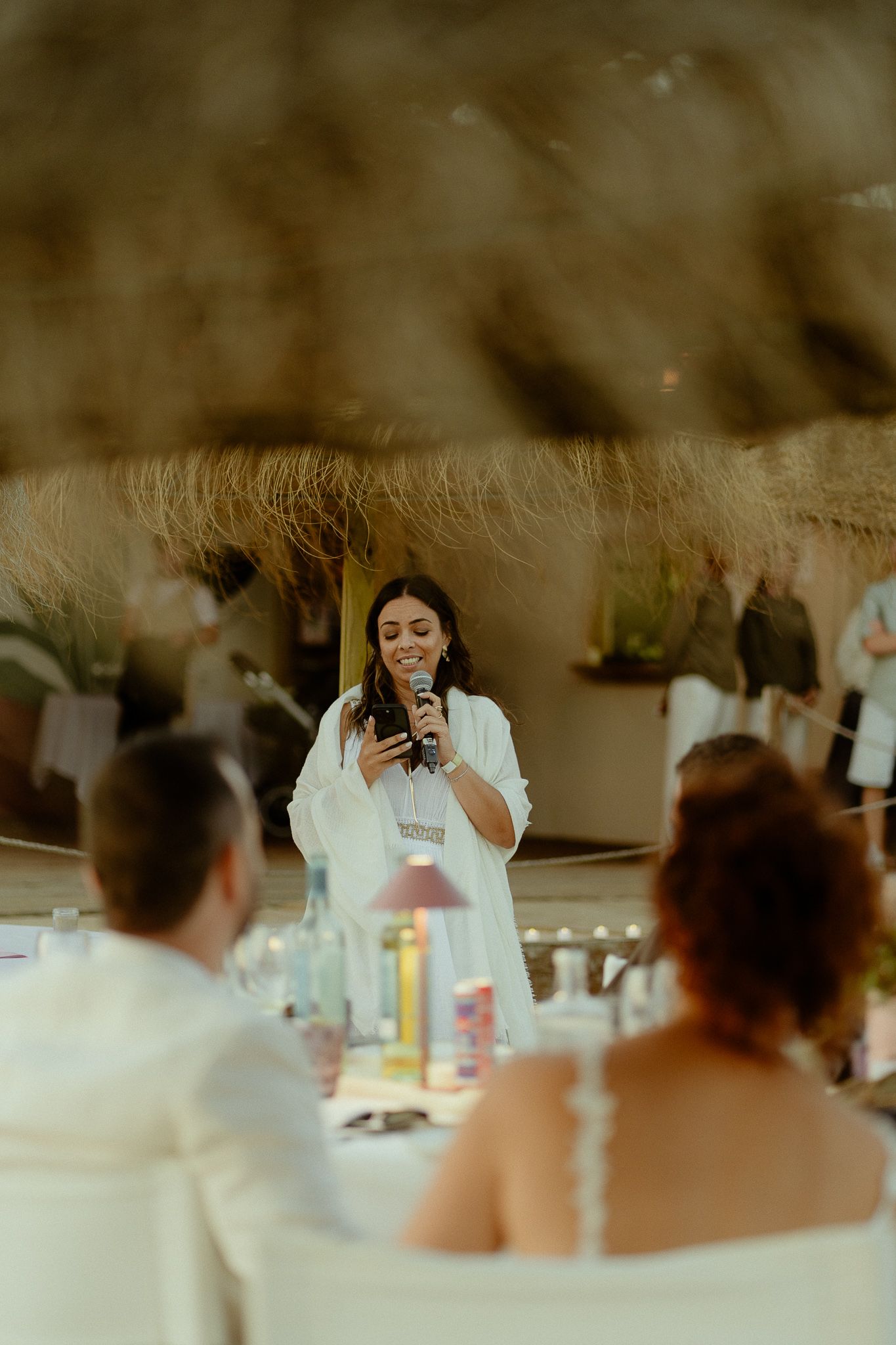 A woman in white gives a heartfelt speech at an elegant outdoor wedding reception while holding a microphone.