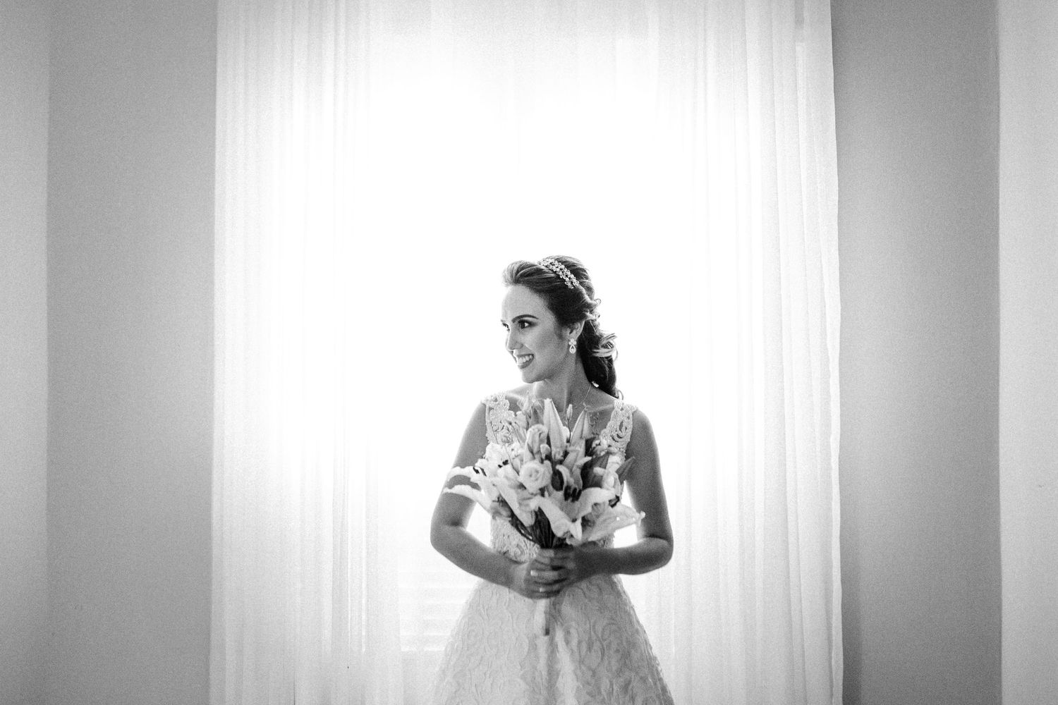 A bride in a white lace wedding dress stands by a window in a timeless black and white portrait.