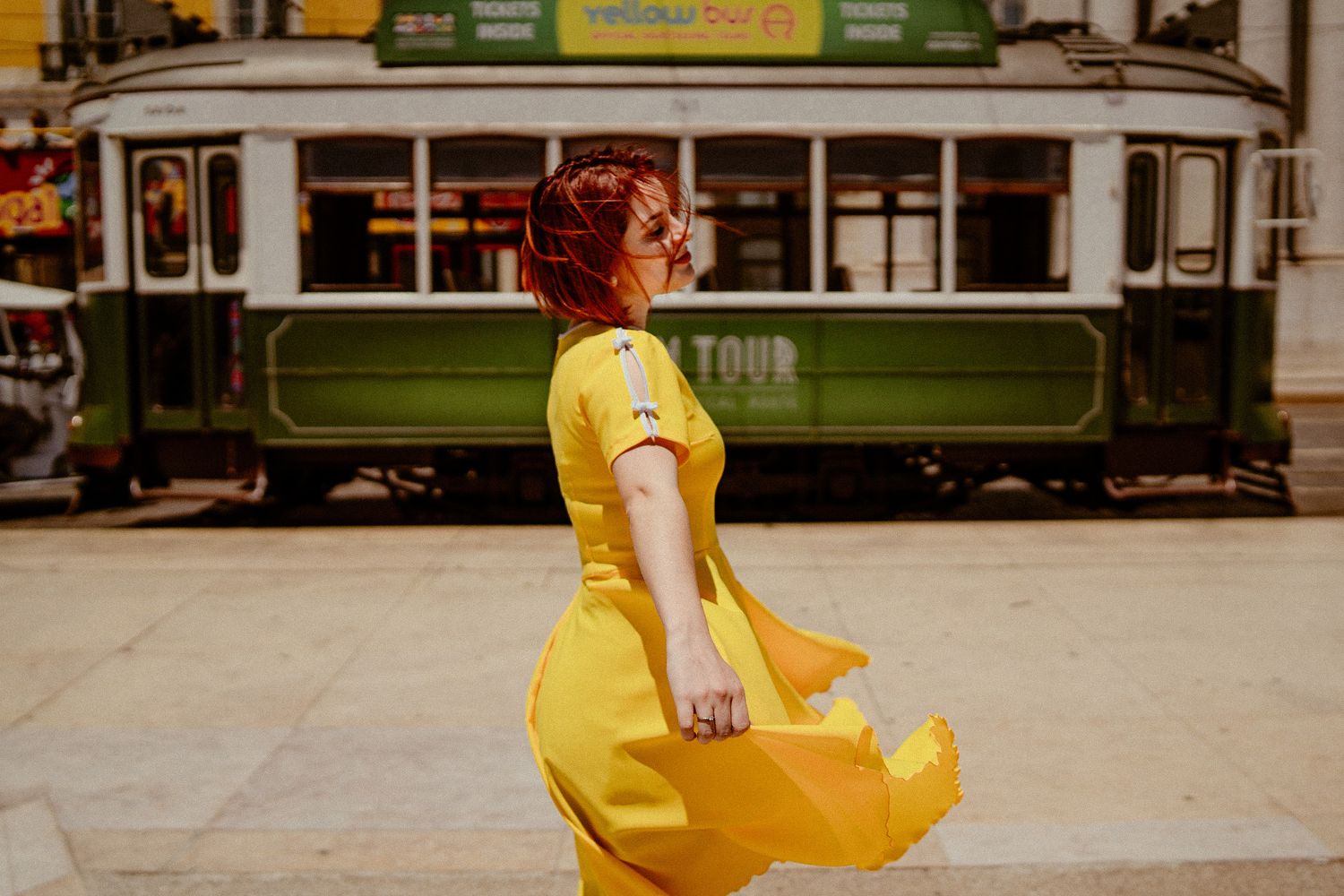 A yellow dress catches the breeze beside a vintage green tram car.