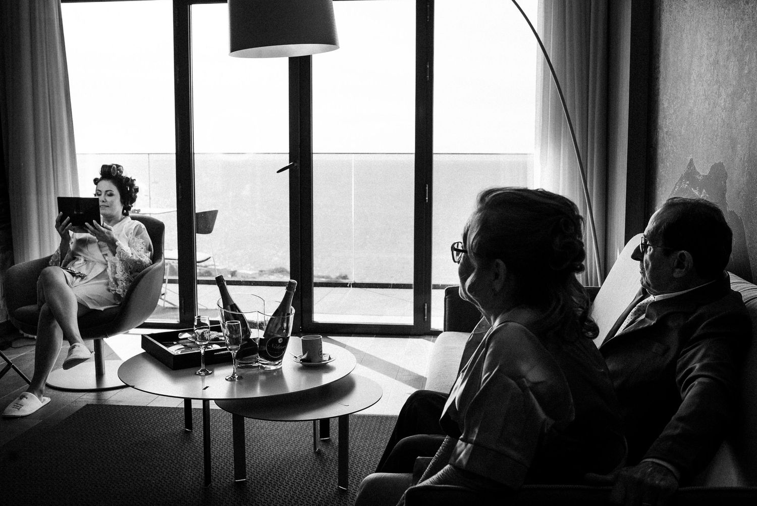 A black and white photograph captures silhouetted figures sitting at a table next to a window with a lamp.