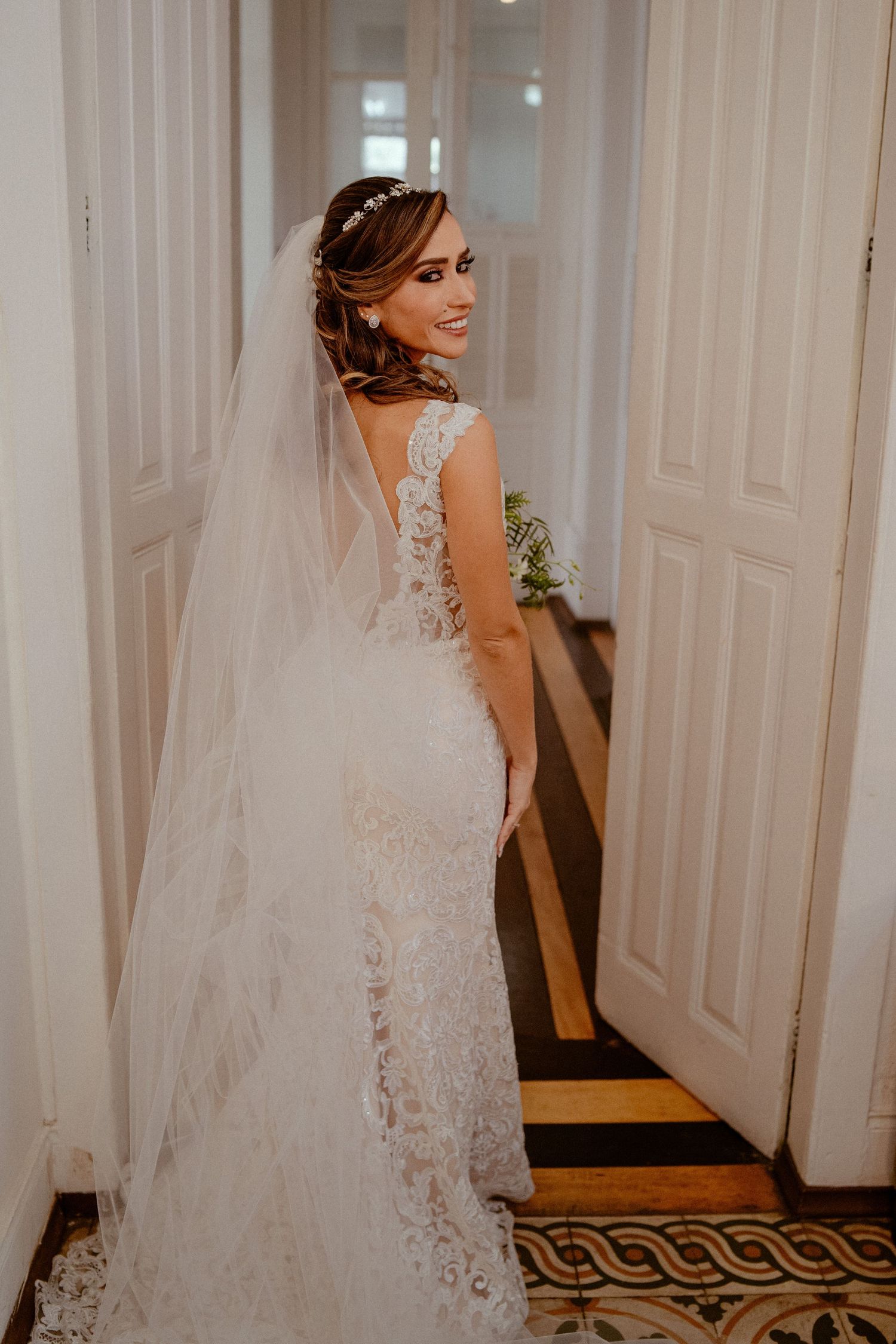 Bride in elegant lace gown and long veil poses gracefully in a hallway with ornate carpet and white doors.