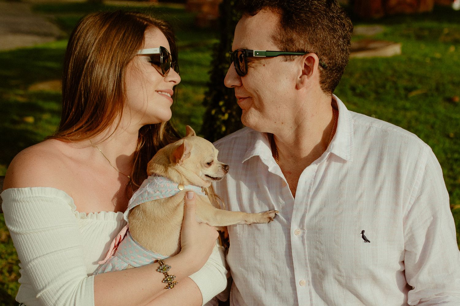A couple in sunglasses shares a tender moment outdoors while holding their small dog in a sunny park setting.