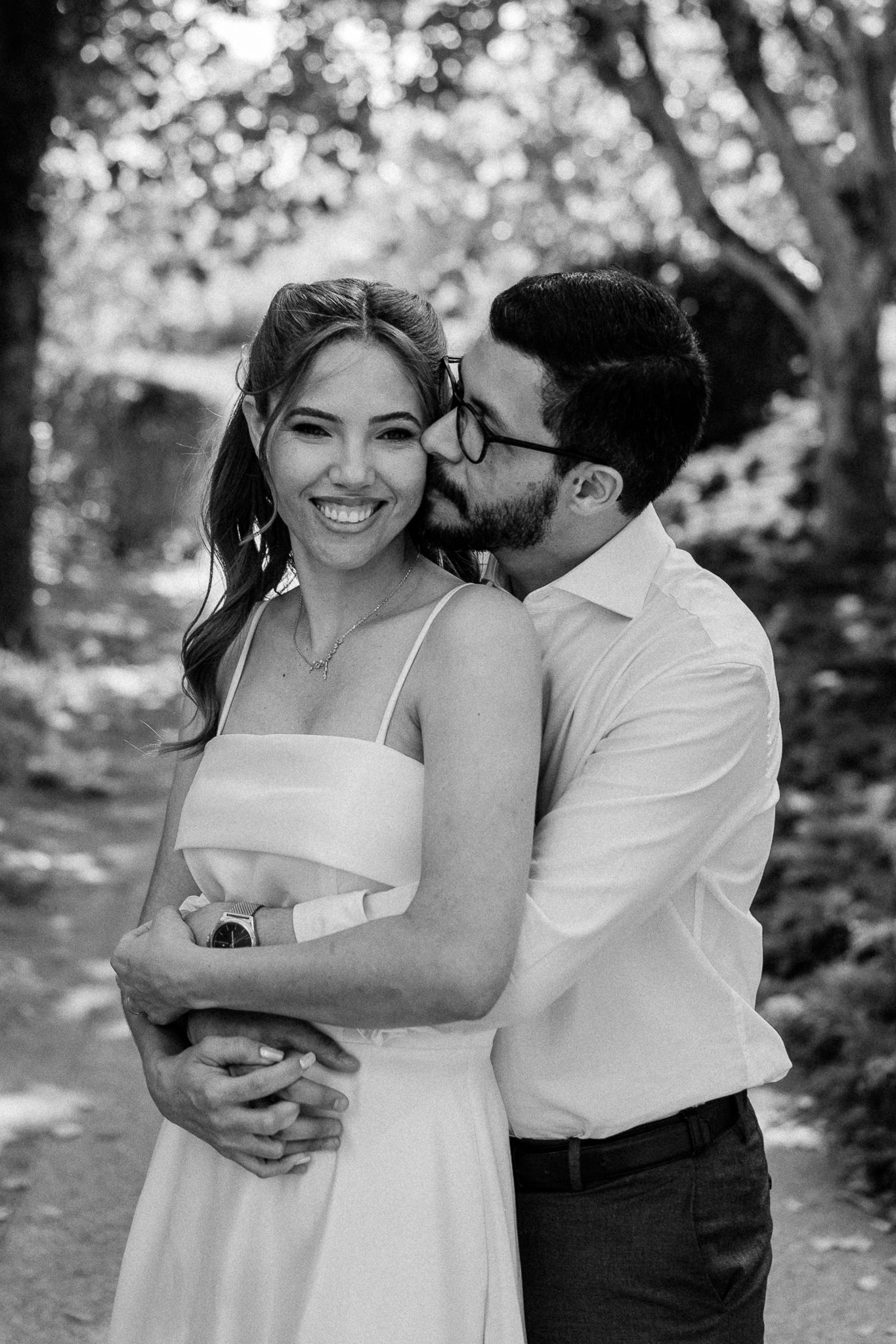 Black and white photo of a smiling woman in white dress being embraced and kissed on cheek by a man outdoors.