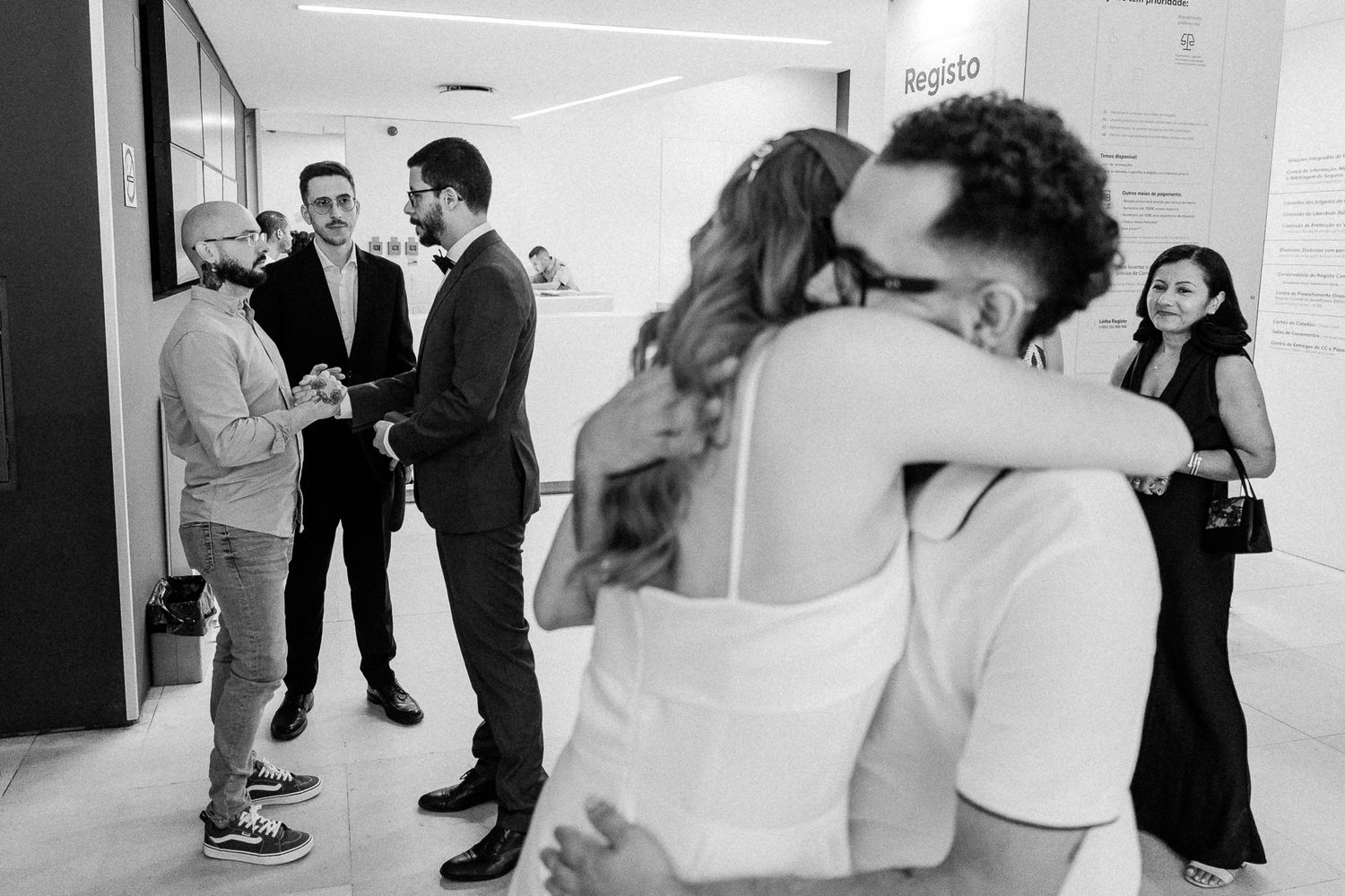 Black and white wedding photo of couple embracing while guests celebrate in background.