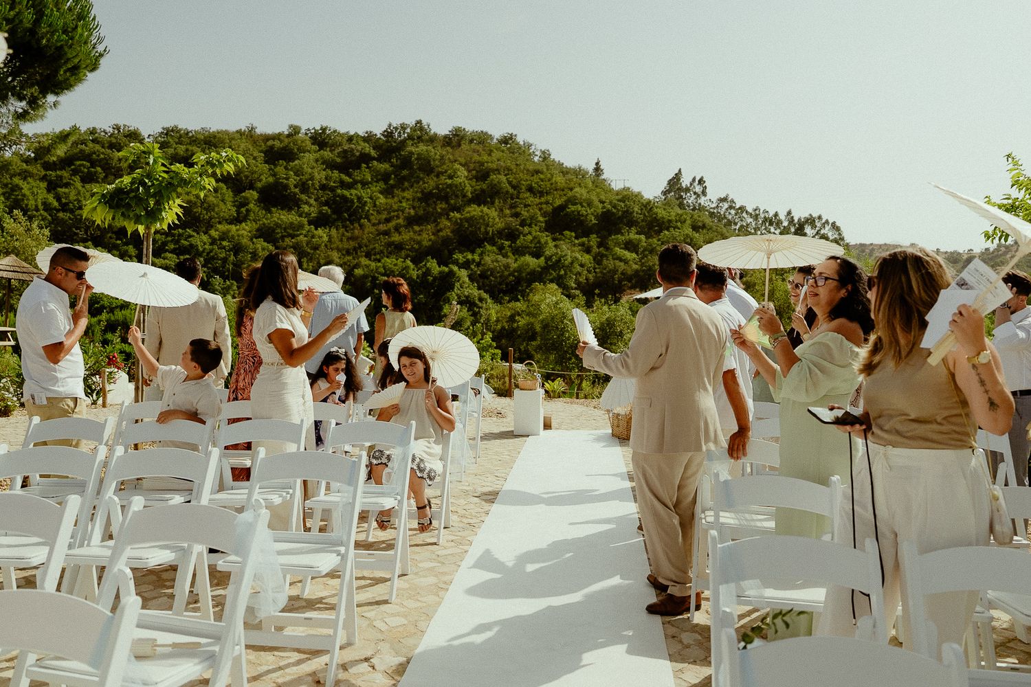 Outdoor wedding ceremony with guests holding white parasols, white chairs, and lush green trees in the background.