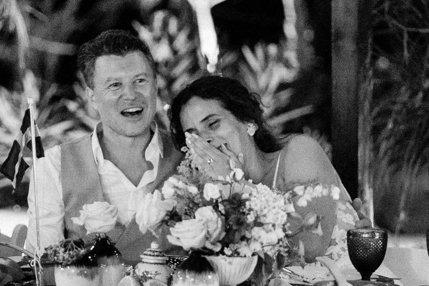 Black and white photo of a laughing couple at their wedding reception table surrounded by floral arrangements.