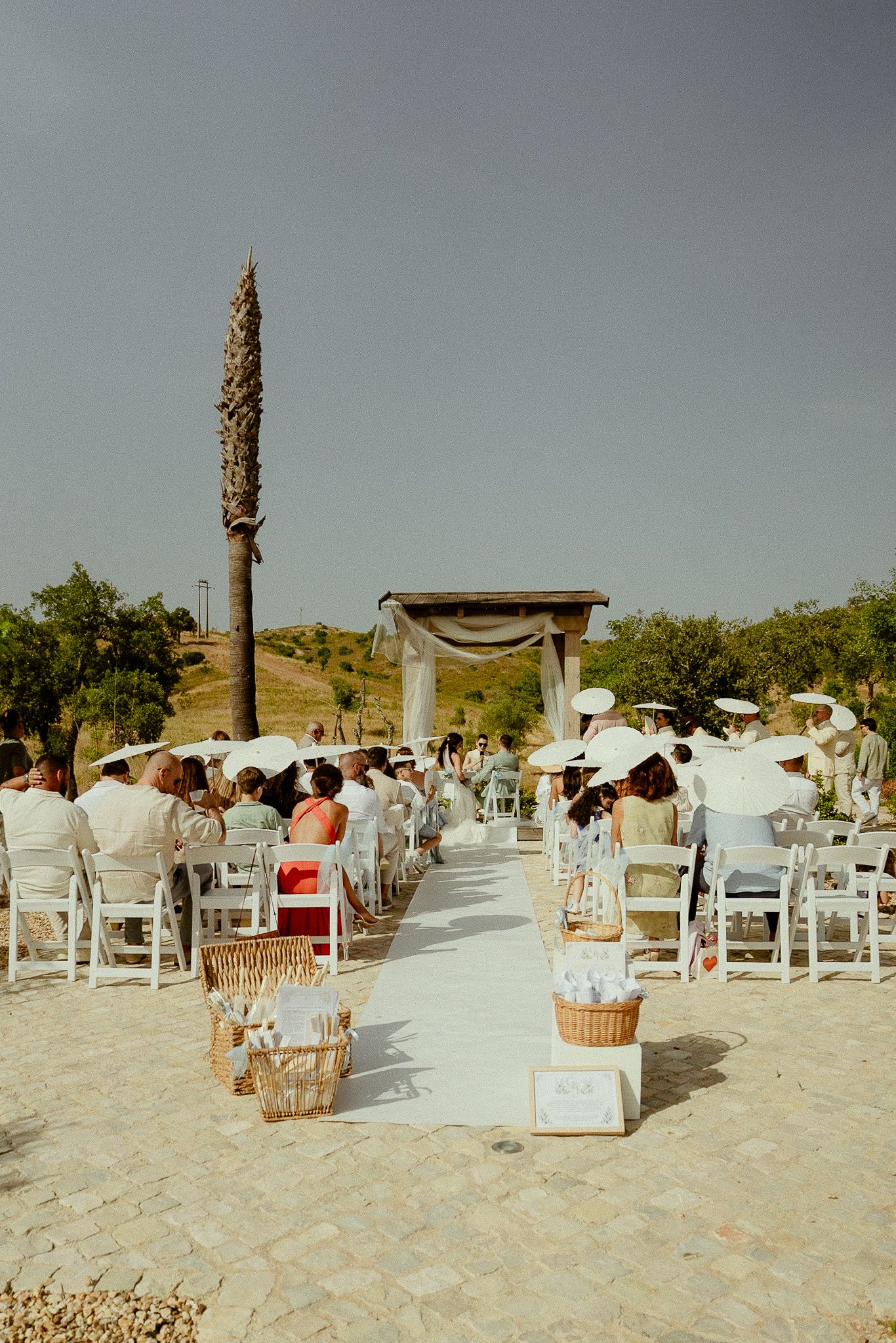 Outdoor wedding ceremony with white chairs, rustic wooden arch, and guests seated under clear blue sky.