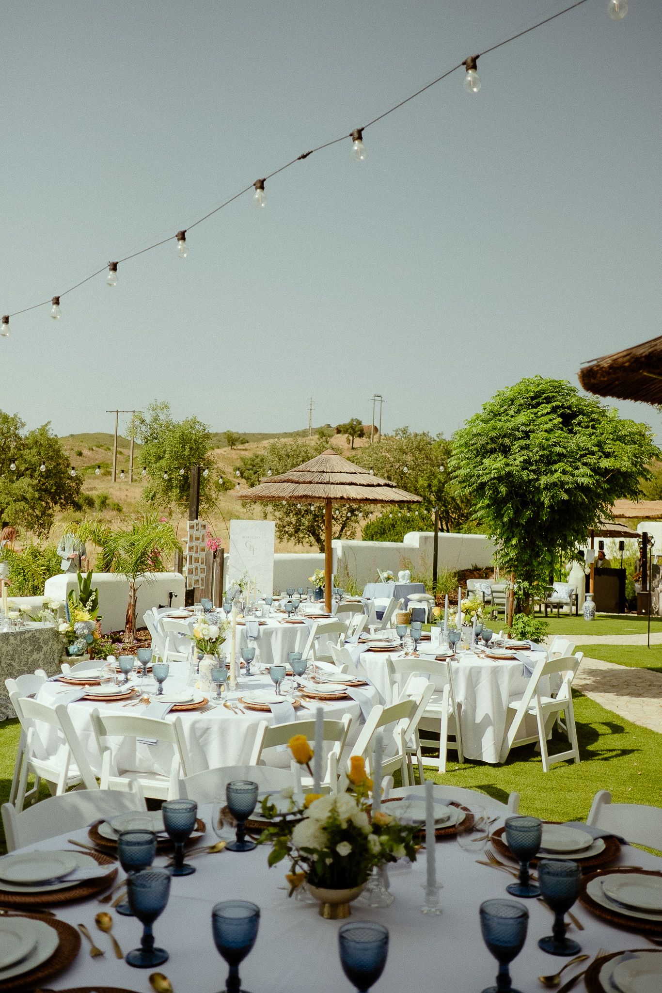 Outdoor wedding reception setup with white round tables, blue glassware, floral centerpieces, string lights, and lush green garden backdrop.