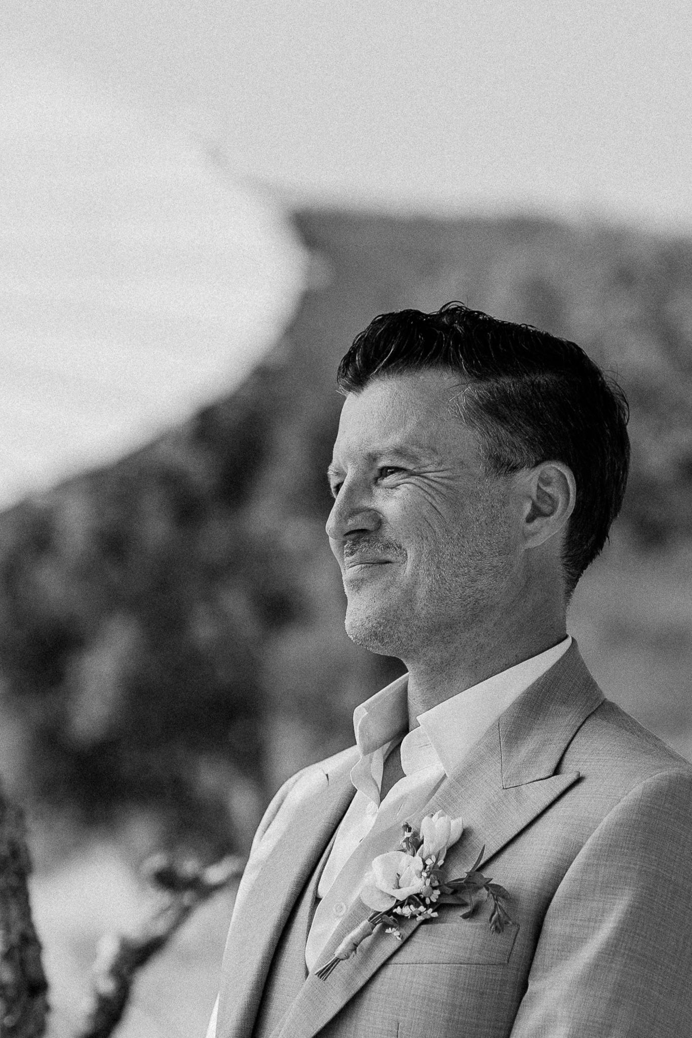 Black and white portrait of a smiling groom in a suit with boutonniere at an outdoor wedding ceremony.