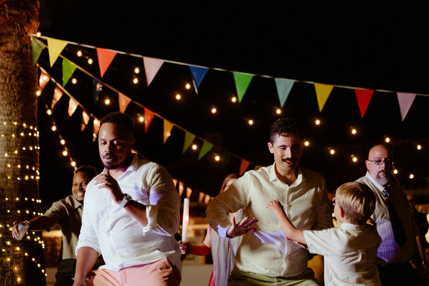 Men and a child dancing outdoors at night under colorful bunting flags and string lights at a festive celebration.
