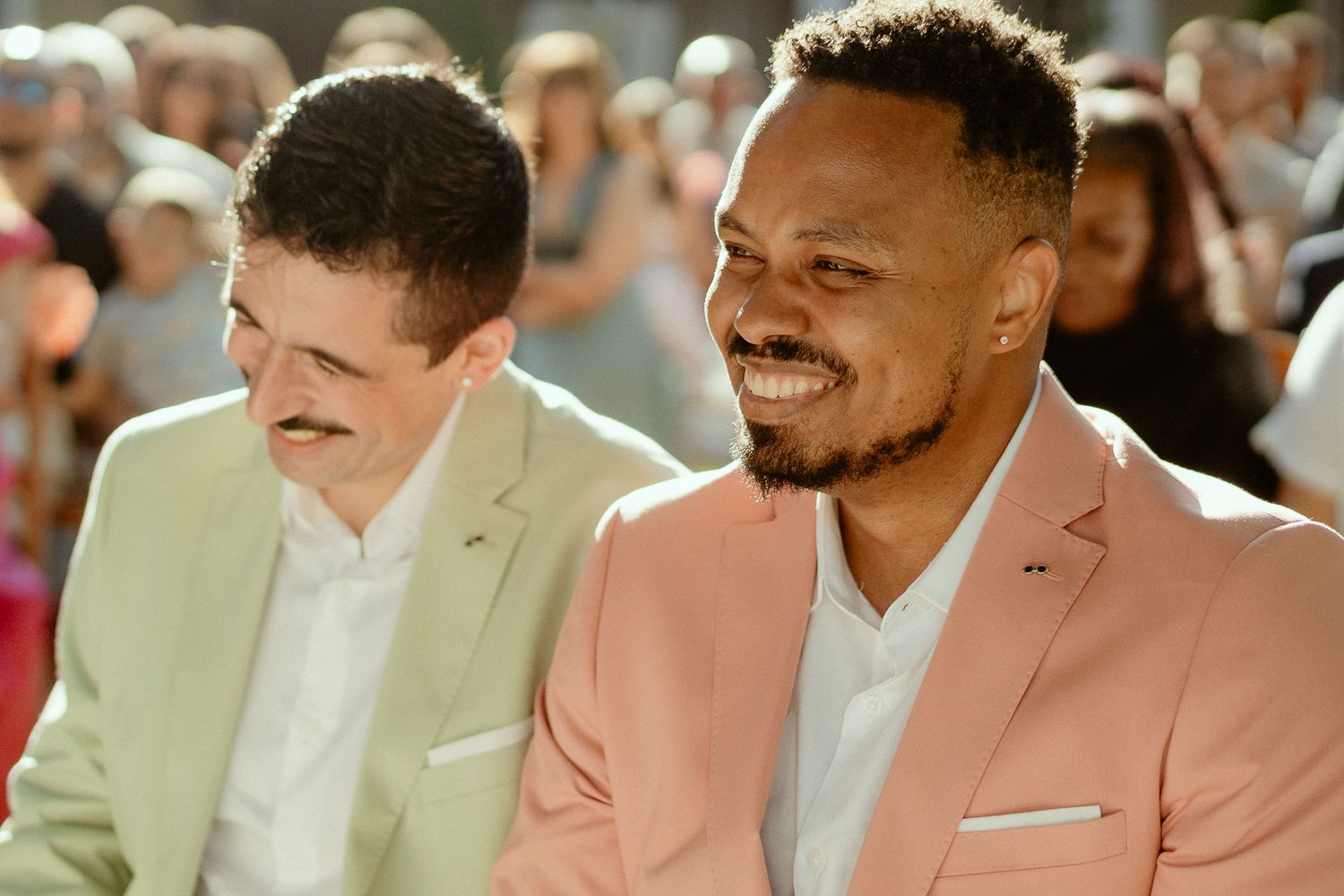 Two men in suits smiling and laughing together at an outdoor event, one in pink blazer.