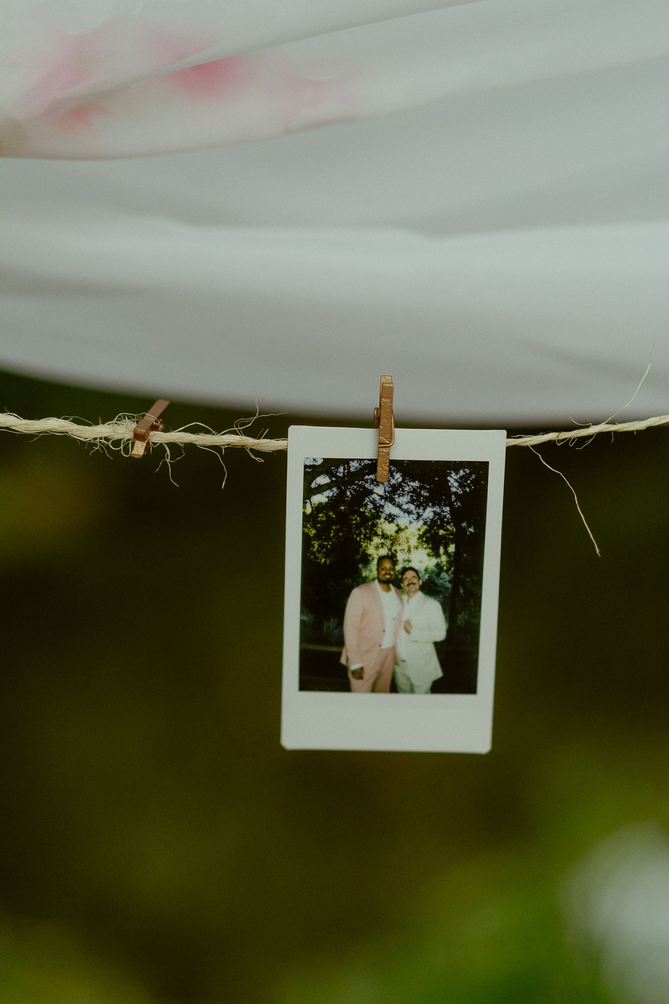 A Polaroid photo of a couple in wedding attire clipped to a twine string outdoors.