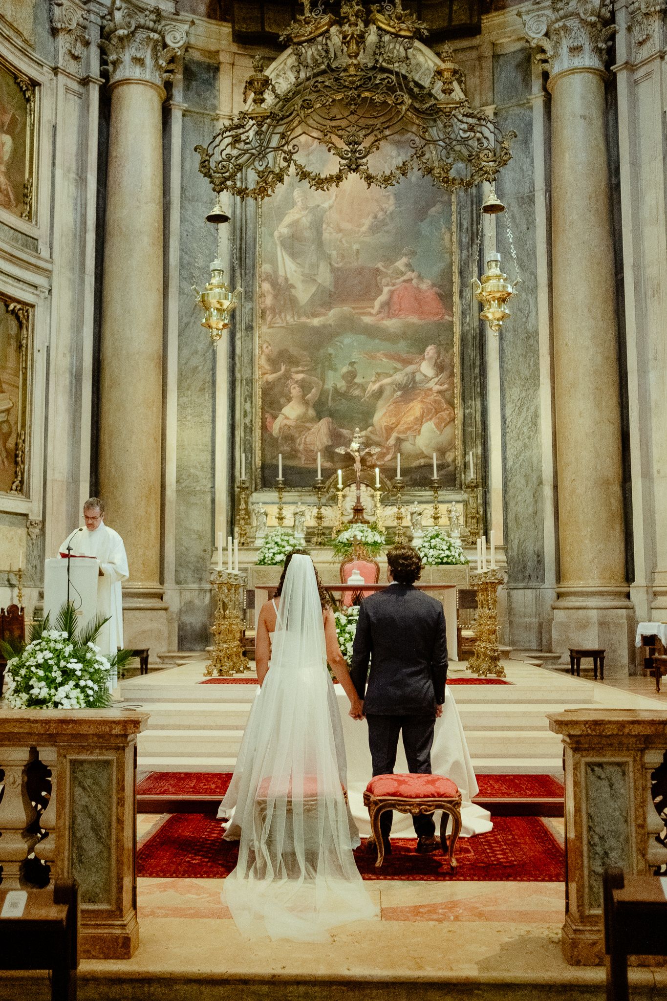 Bride and groom standing at ornate church altar during wedding ceremony with large baroque painting backdrop.
