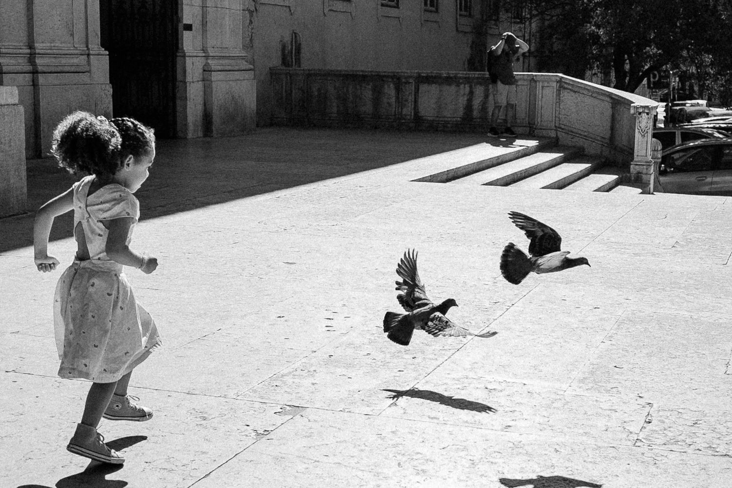 Black and white photo of a young girl in a dress chasing pigeons on a sunny plaza.