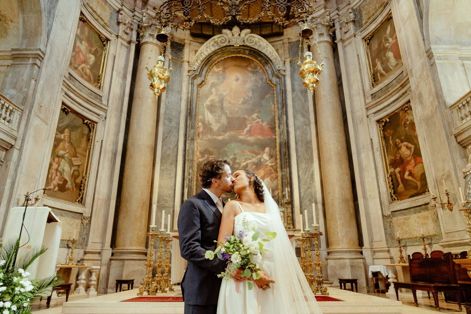 Bride and groom share a kiss at the altar of an ornate baroque church with grand paintings and golden columns.