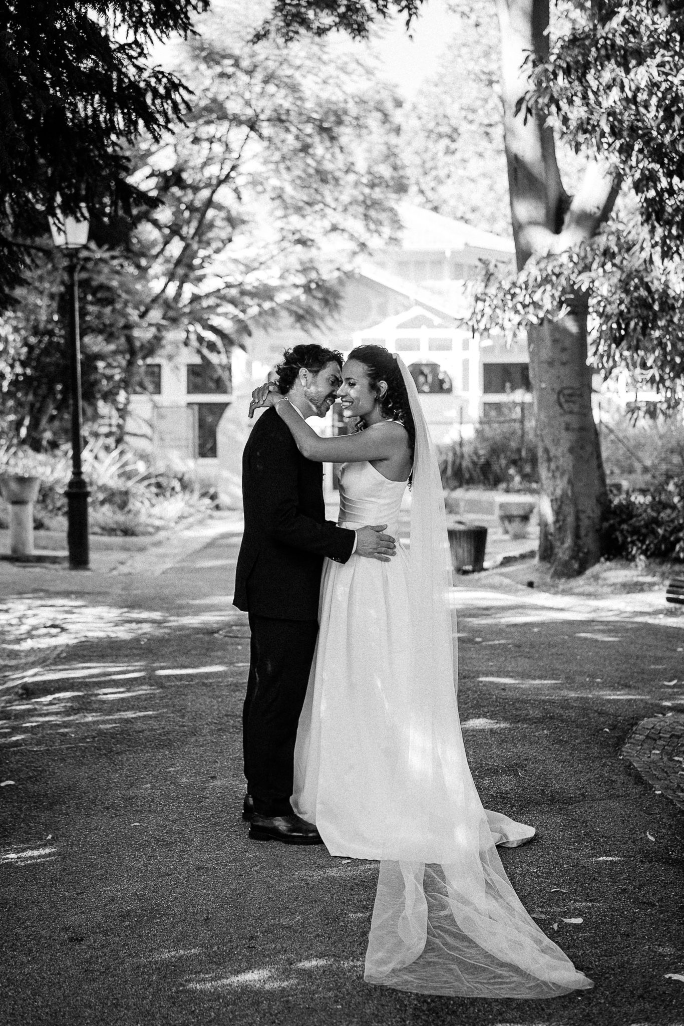 Black and white photo of a bride and groom embracing outdoors, sharing a romantic moment in a tree-lined park setting.