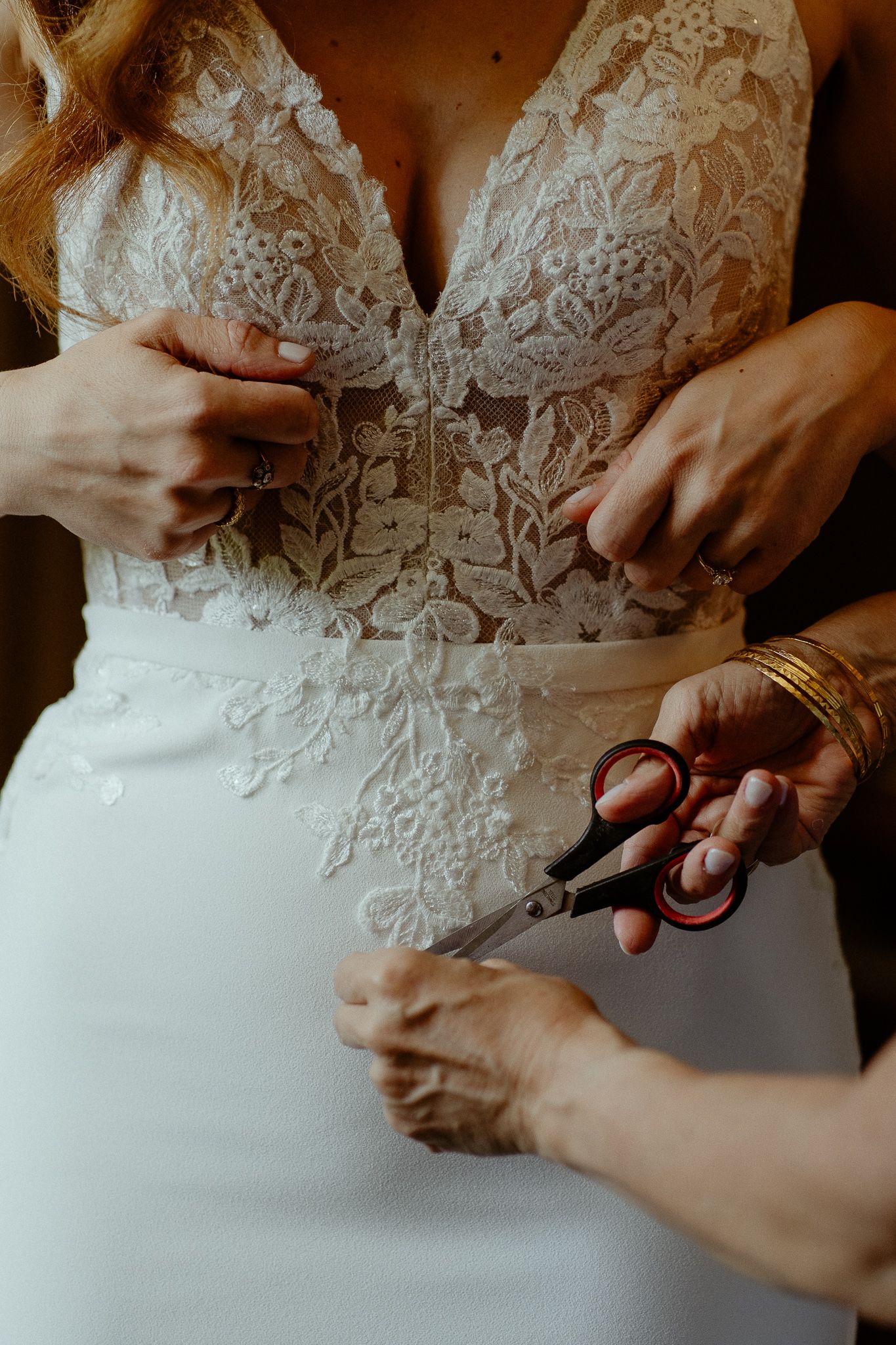 A bride in a lace wedding gown being fitted with scissors adjusting the dress waist by helping hands.