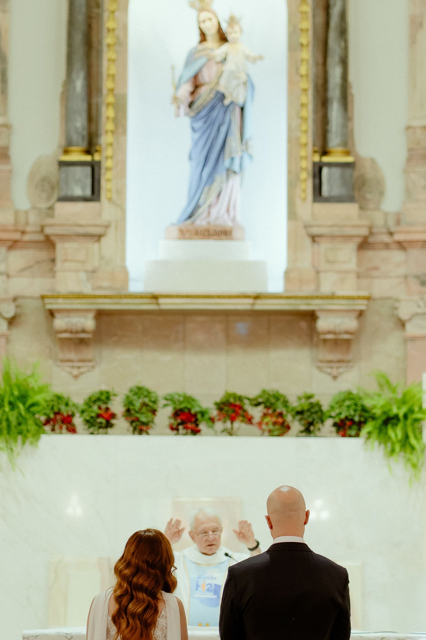 Couple stands before a priest at a Catholic church altar with a Virgin Mary statue during a wedding ceremony.
