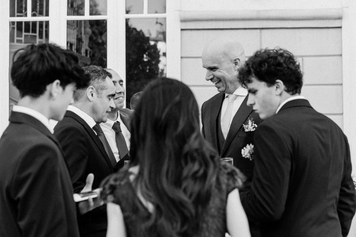 Black and white photo of elegantly dressed men in suits conversing and smiling at a formal wedding reception.