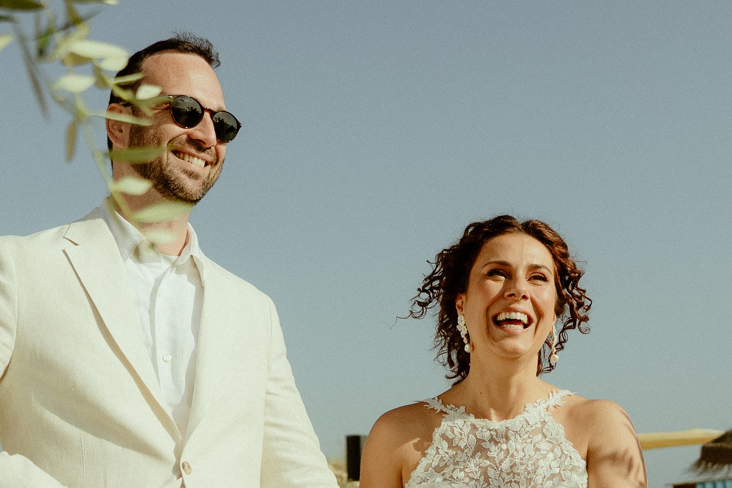 Happy bride and groom laughing outdoors on a sunny day, groom in white suit with sunglasses, bride in beaded halter gown.