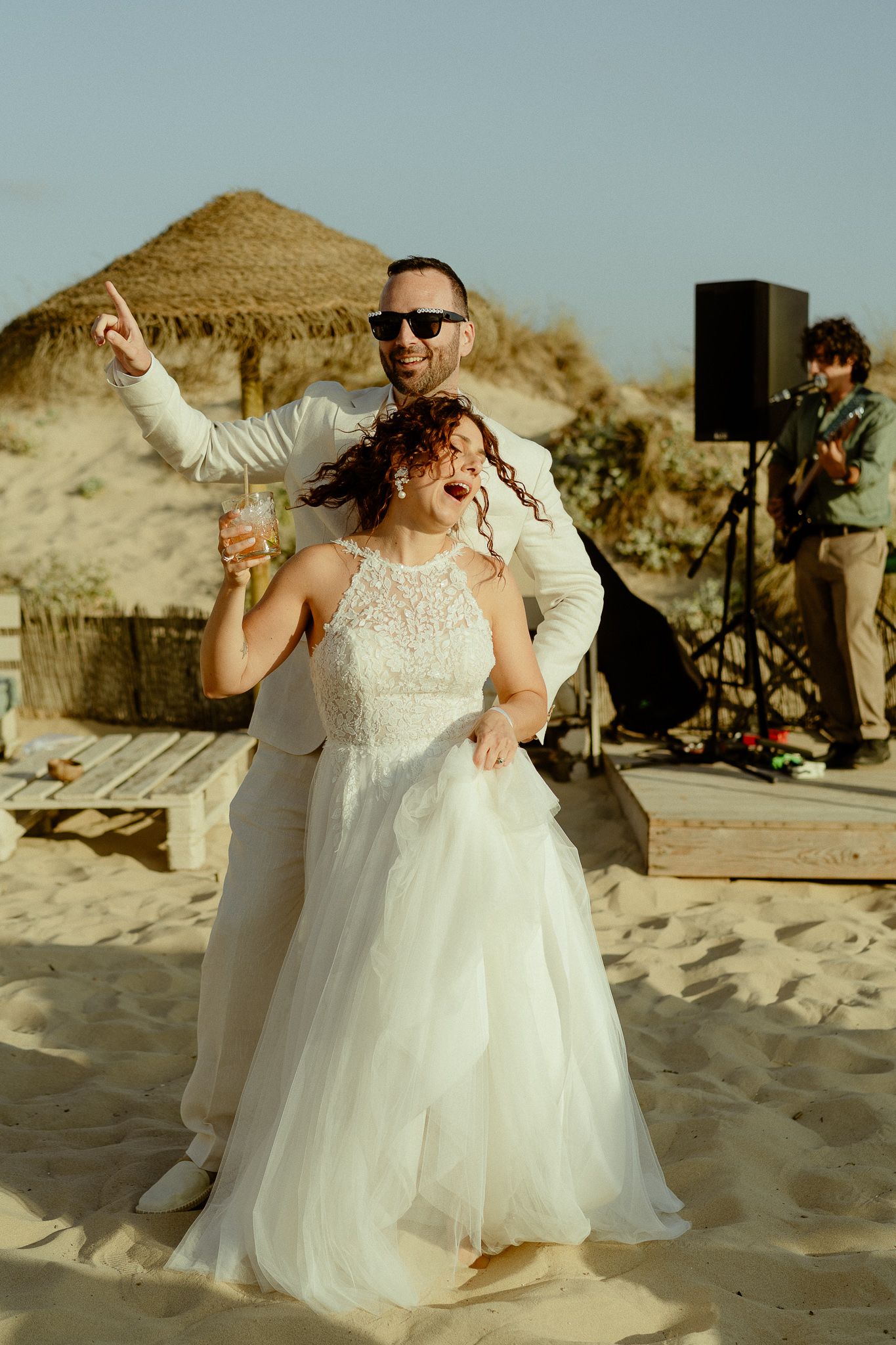 Bride and groom dancing joyfully on sandy beach at outdoor wedding reception with live musician performing.