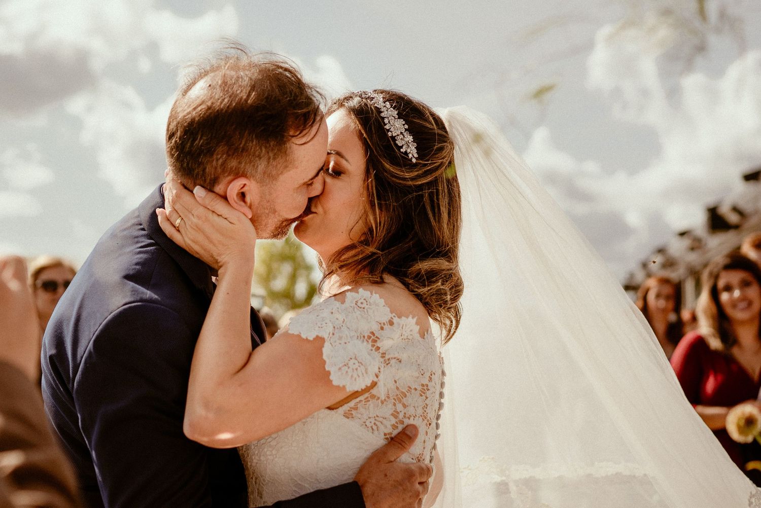Married couple shares a passionate kiss during outdoor wedding ceremony while guests look on in background.