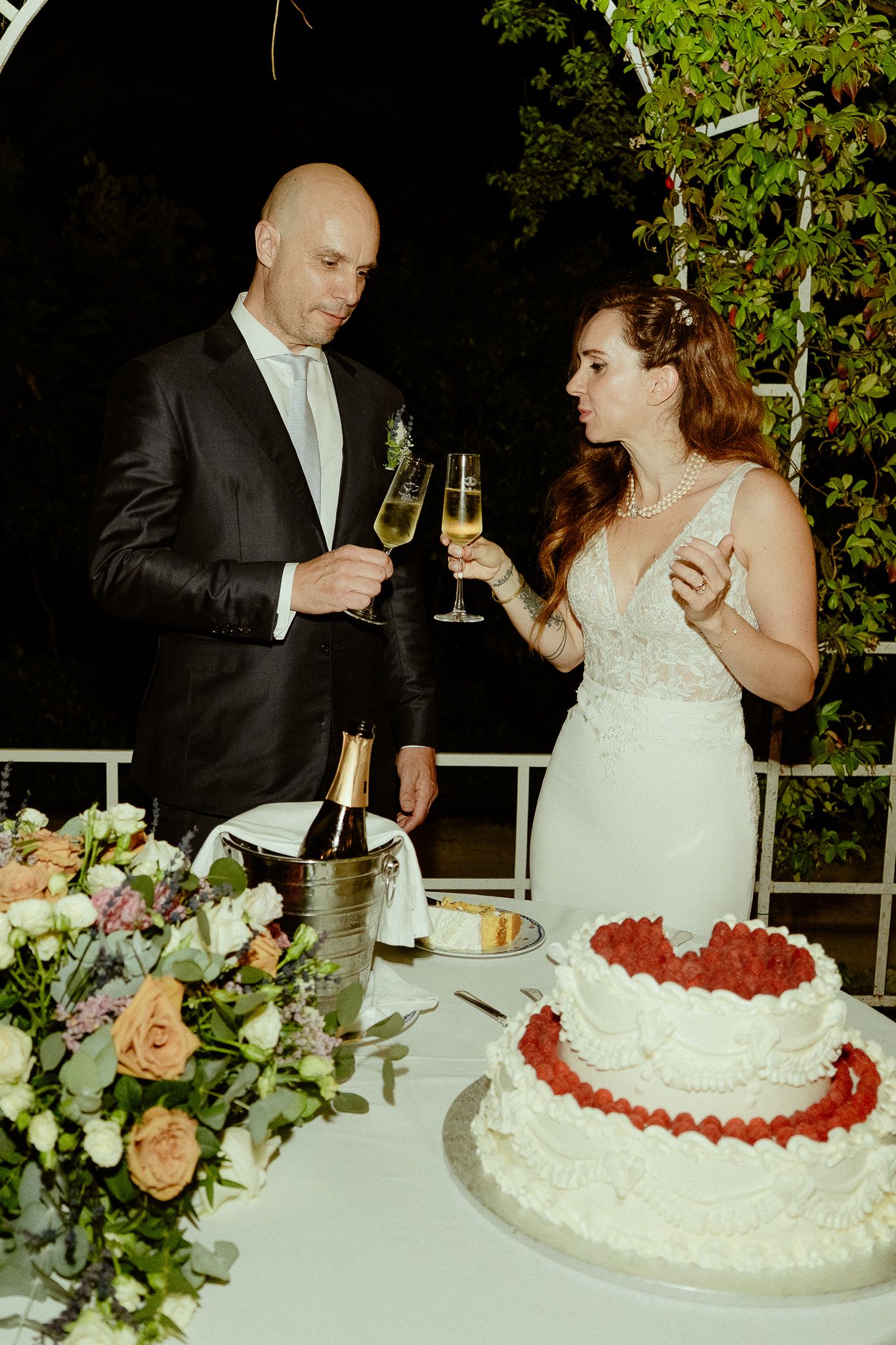 Bride and groom toasting with champagne glasses beside a two-tier white wedding cake decorated with fresh strawberries.