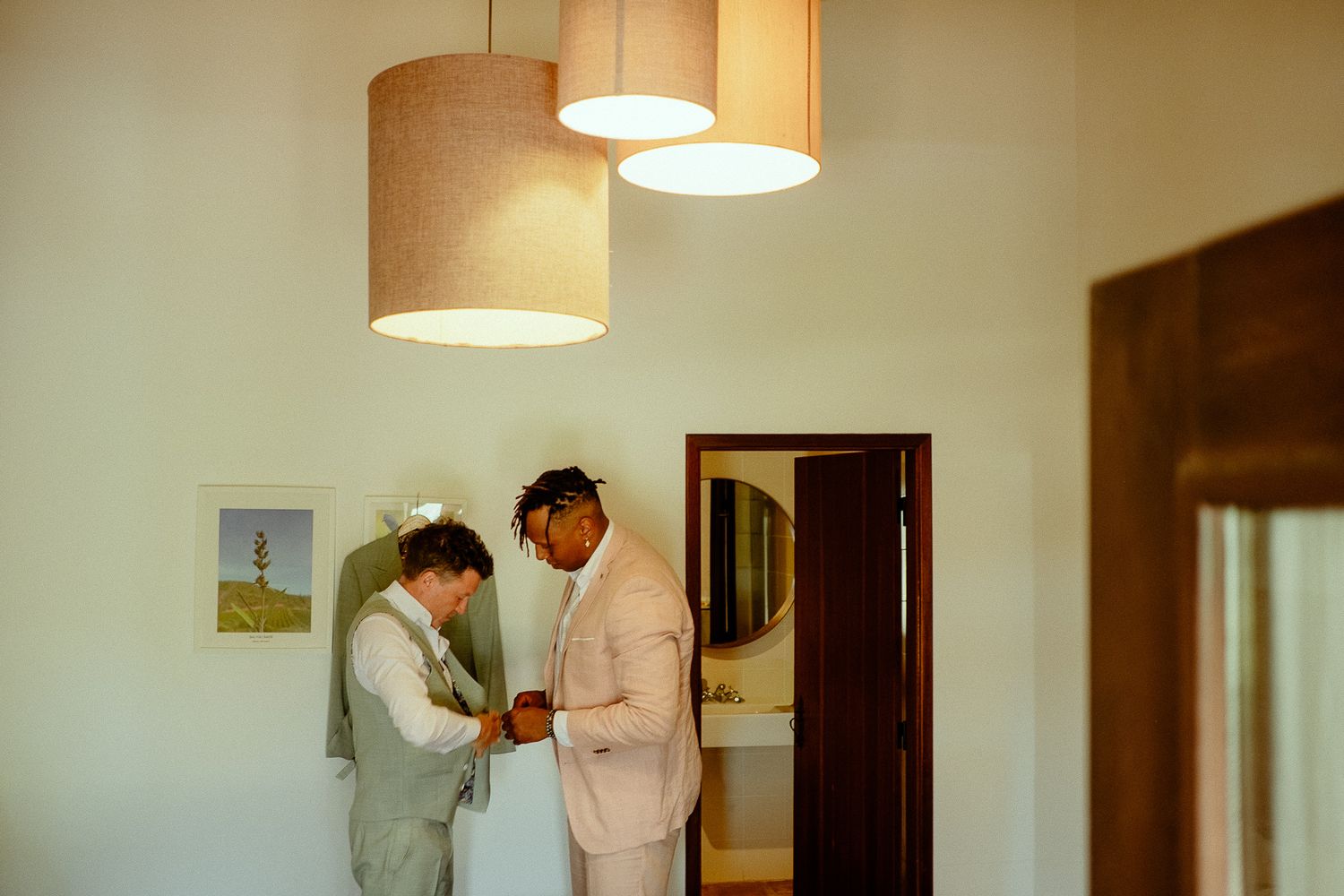 Two grooms helping each other dress in a bright room with pendant lights on their wedding day.