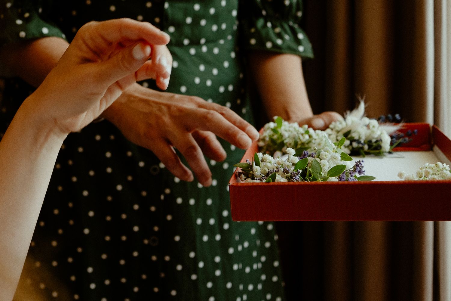 Hands exchanging floral wedding accessories on a red tray, with a woman in a green polka dot dress.