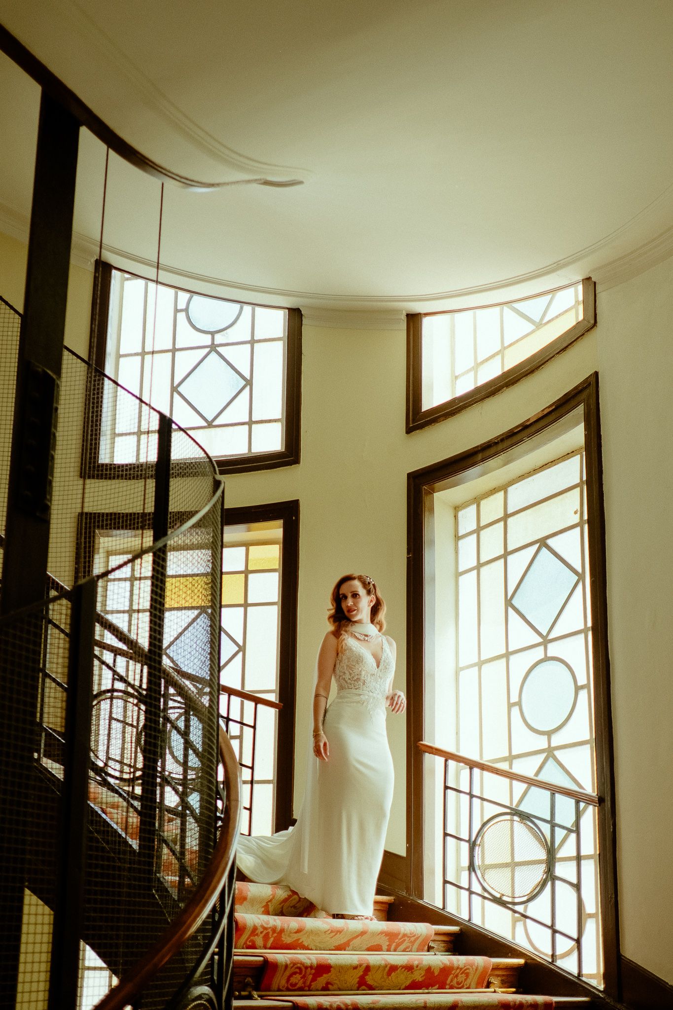Bride in white gown standing on ornate staircase with stained glass windows in elegant venue.