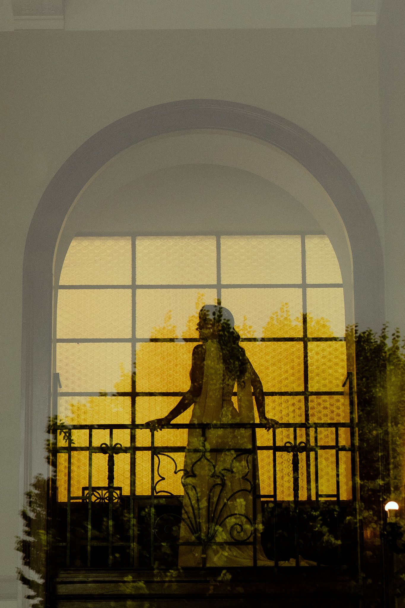 Silhouette of a woman in a white dress standing on an ornate balcony framed by an arched window with golden light.