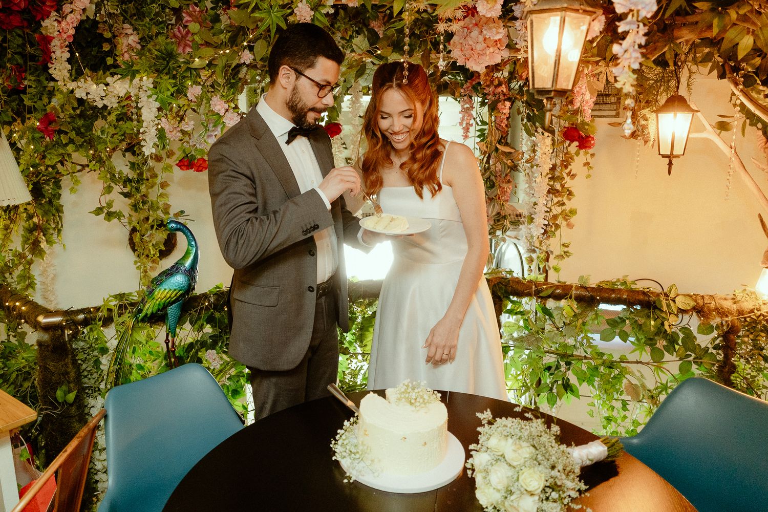 Bride and groom cutting white wedding cake surrounded by lush floral greenery decor at elegant garden-themed reception.