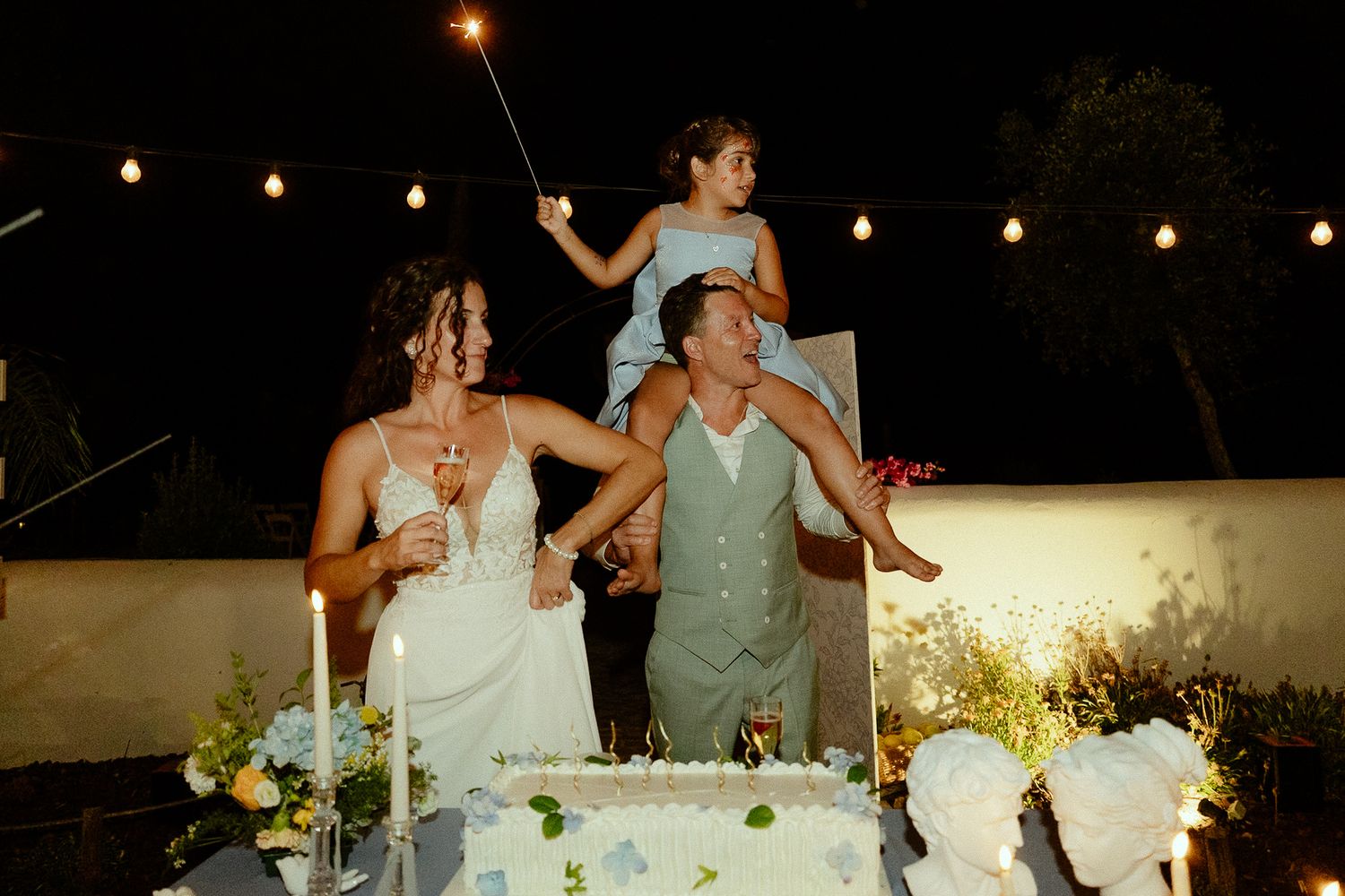 Bride and groom cutting wedding cake at night with flower girl on shoulders holding sparkler.