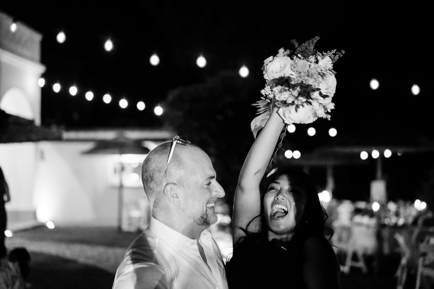 Black and white photo of a joyful couple at a wedding reception, bride raising bouquet under string lights at night.
